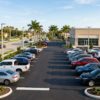 A clean, well-maintained outdoor used car dealership lot on a wide commercial boulevard in Florida. Several clean used vehicles (mix of sedans, SUVs, trucks) of different colors parked in neat rows. The lot is tidy with fresh asphalt and painted lines. Background: palm trees, a wide road with a center median, blue sky with light clouds, warm afternoon sunlight. The scene is inviting and professional — not cluttered, not flashy. NO dealership signs, NO business names, NO banners, NO manufacturer badges, NO license plates. The building behind is a clean modern commercial structure with NO text. Photorealistic, editorial automotive photography. 16:9, 1200×675px minimum.